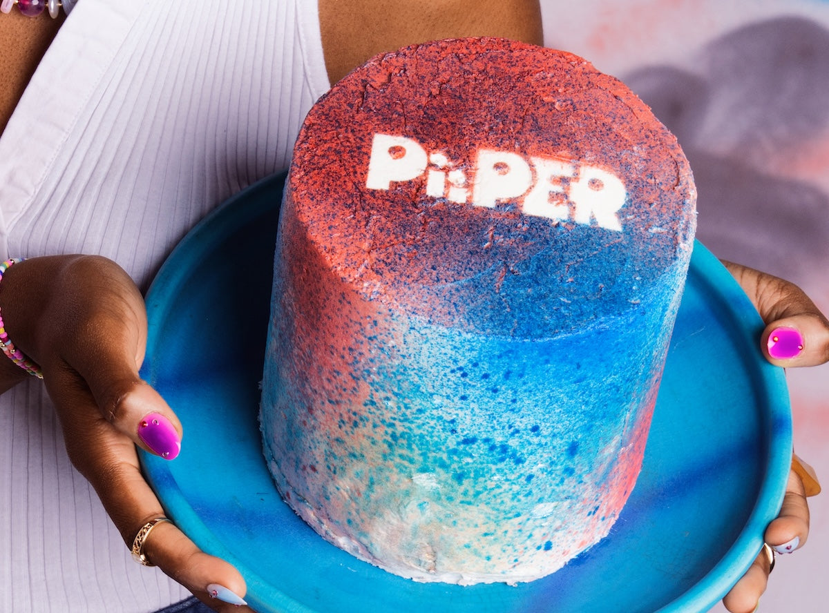 Person holds a blue plate with a colorful cake decorated in blue and pink PiiPER lettering