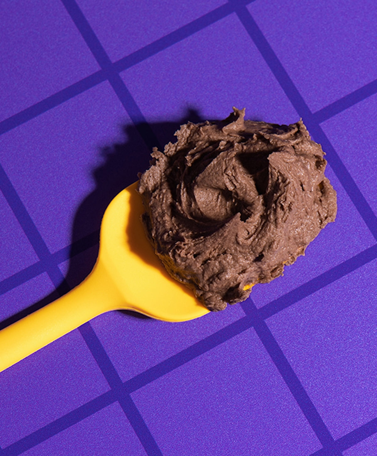 Close-up of Brownie Batter frosting on a yellow spoon showing thick, fluffy texture on a purple geometric background