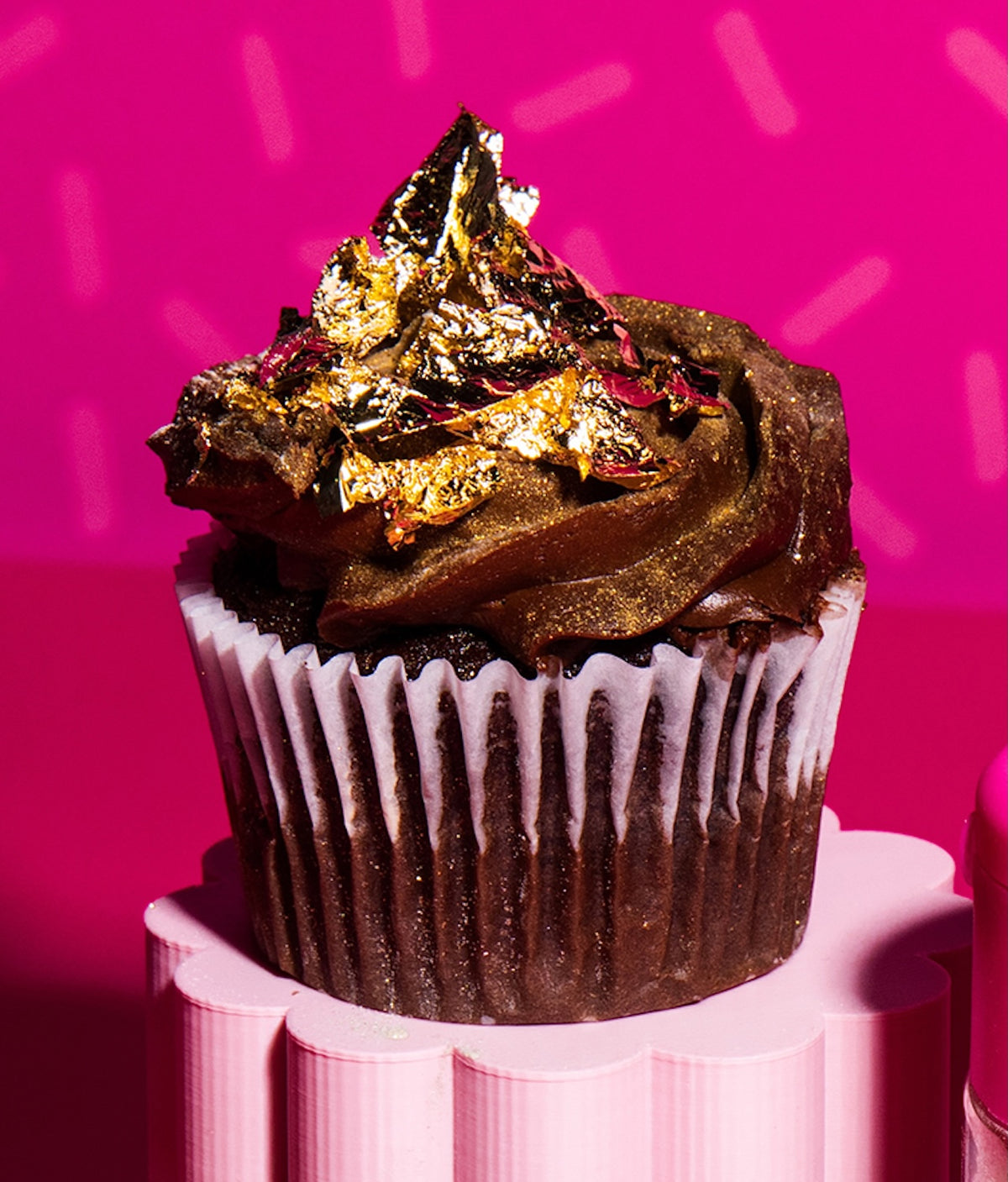 Gold decorative flakes decorate a chocolate cupcake against a bright pink background