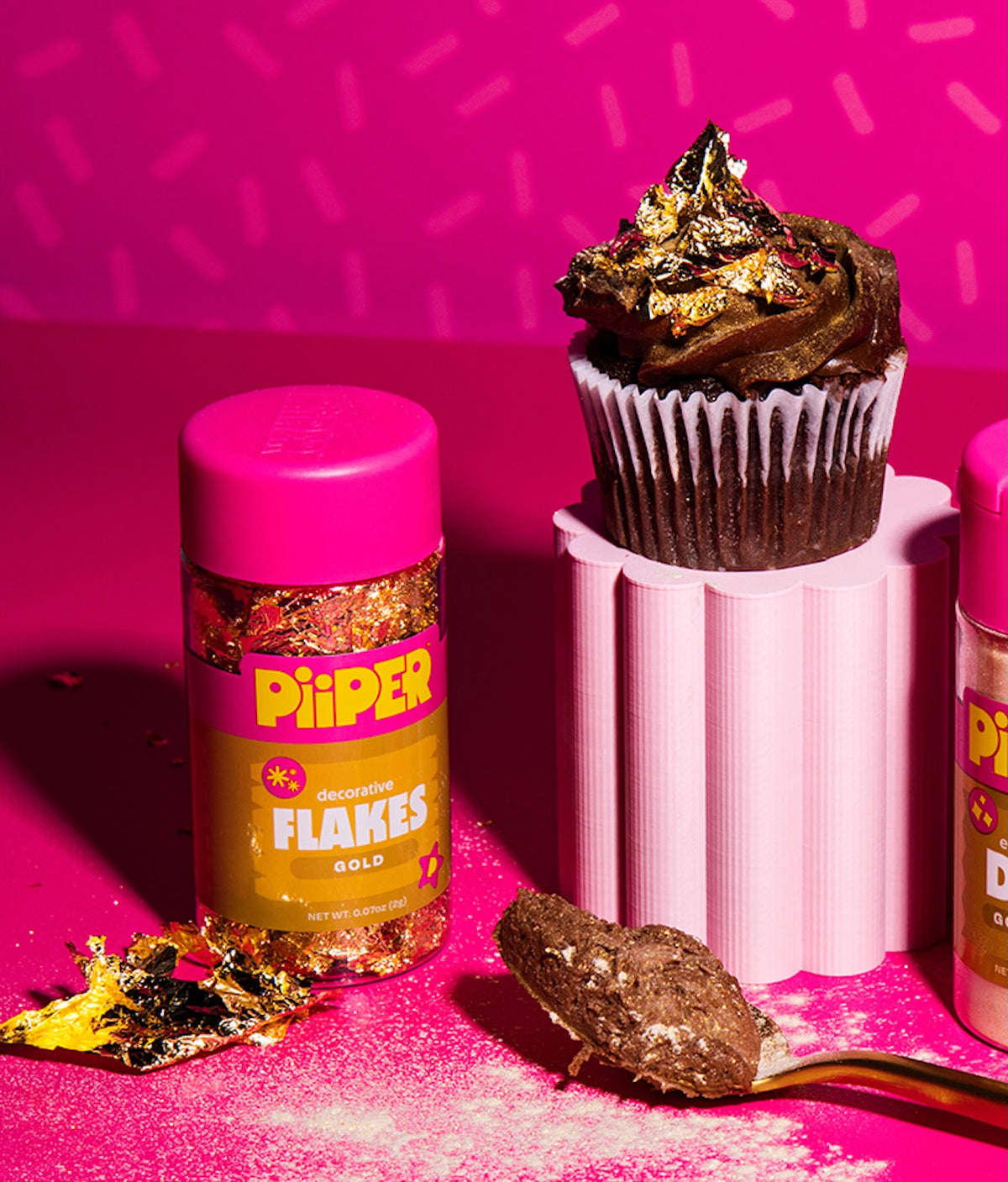 Front view of PiiPER Decorative Gold Leaf Flakes jar showcasing gold flakes on a cupcake against a bright pink background