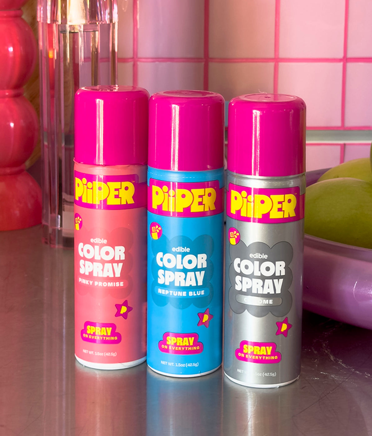 Front view of PiiPER Edible Color Spray in Chrome showing silver packaging beside Pinky Promise and Neptune Blue cans on a reflective counter