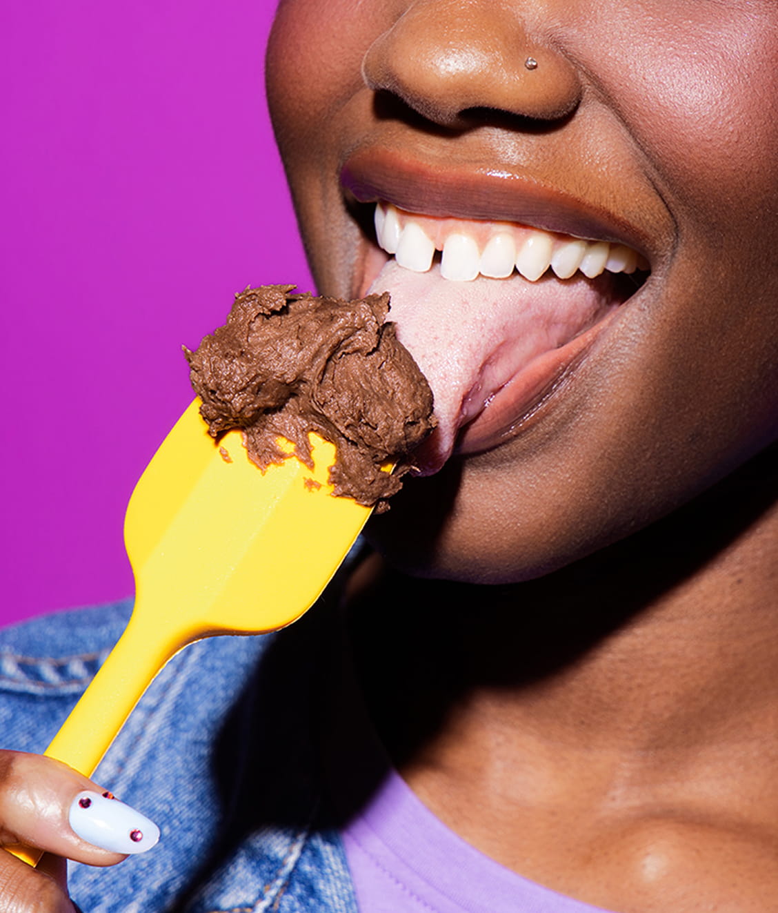 Person eating Brownie Batter frosting from a yellow spatula against a purple background
