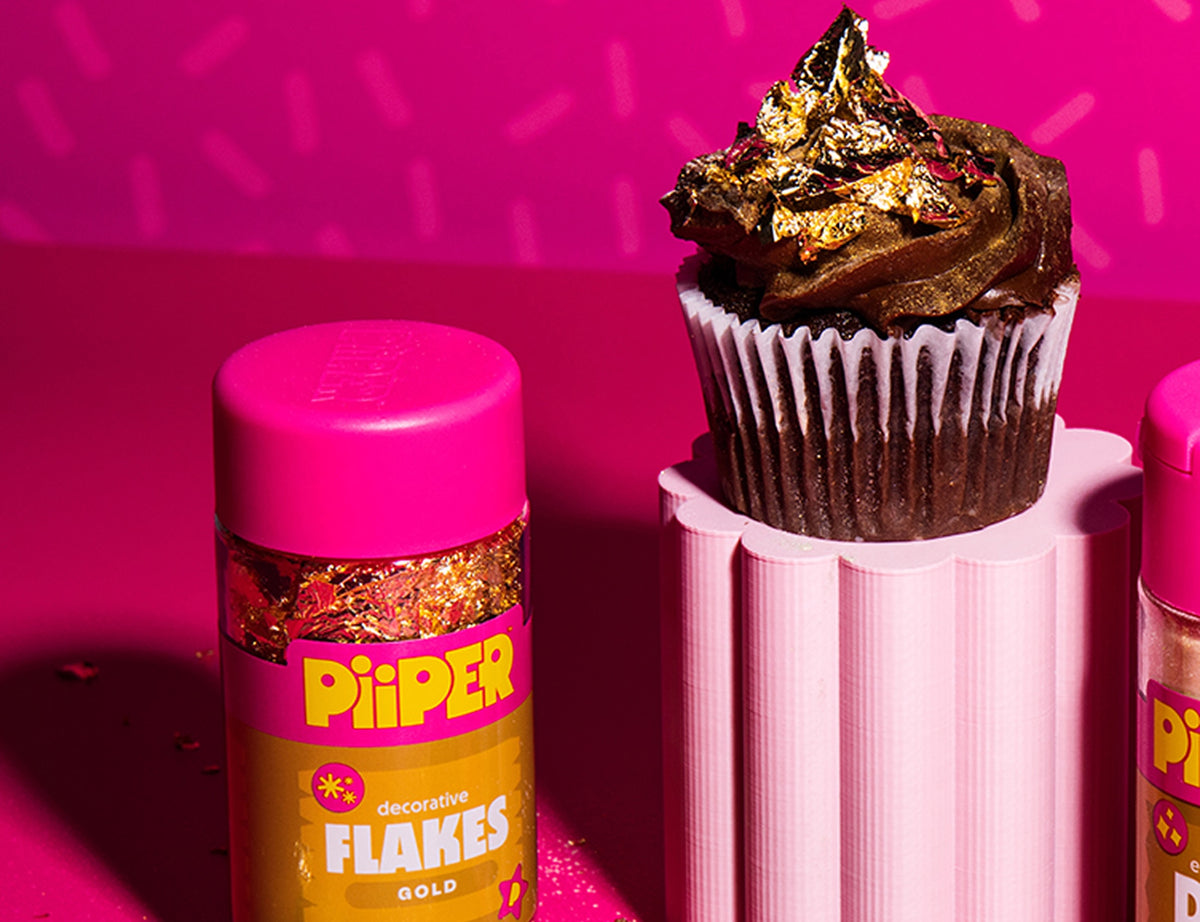 Gold decorative flakes topping a chocolate cupcake beside a PiiPER container on a bright pink background