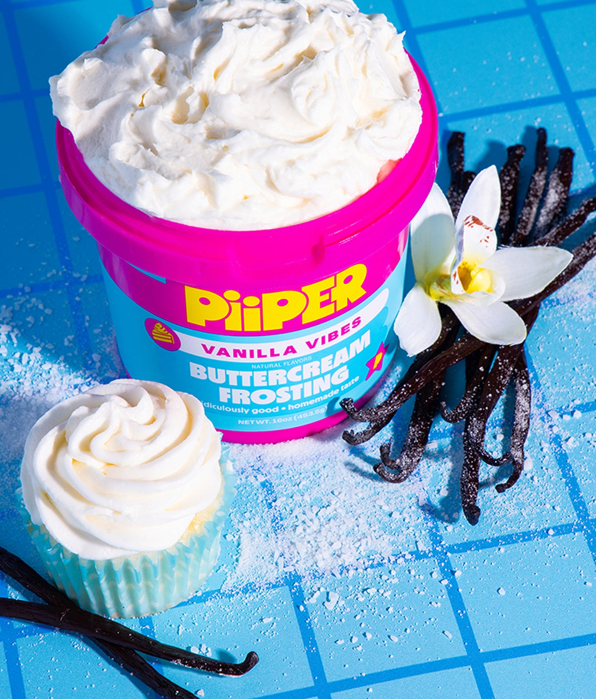 Front view of PiiPER Vanilla Vibes buttercream frosting tub with swirled frosting, cupcake, and vanilla beans on a blue background