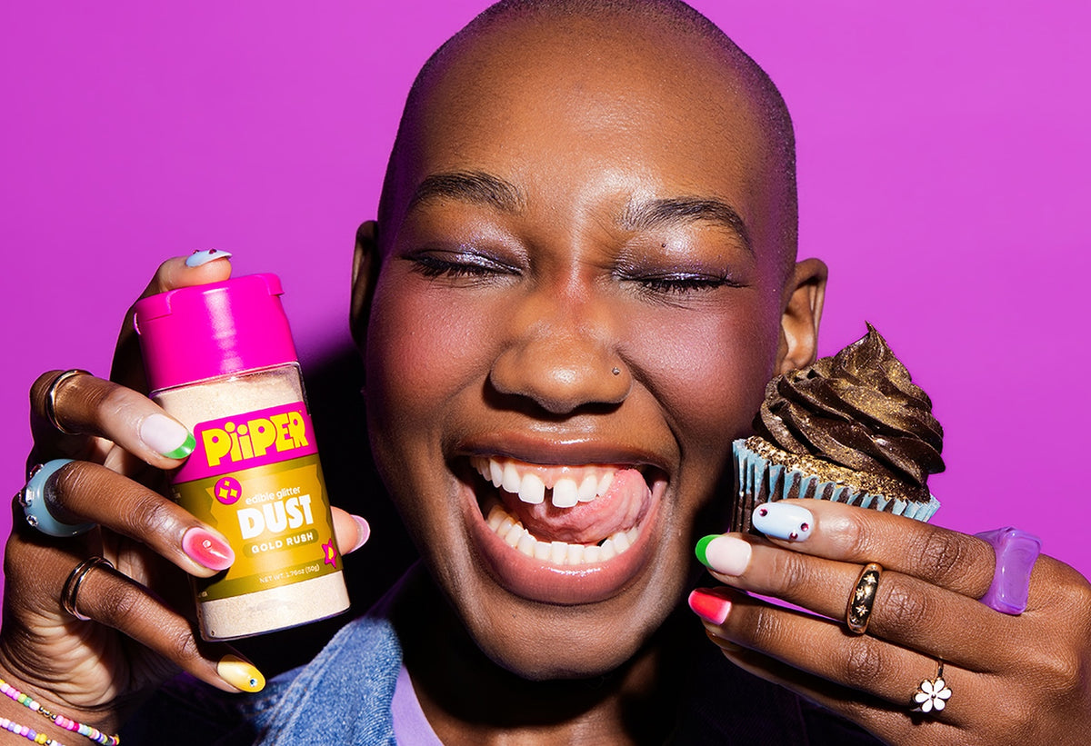 Person smiling while holding PiiPER Gold Rush edible glitter dust and a chocolate-frosted cupcake against a bright purple background